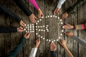 Teamwork and cooperation concept - group of twelve people, male and female, assembling a light bulb shape with blank puzzle pieces on a rustic textured desk, top view. Teamwork and cooperation concept.