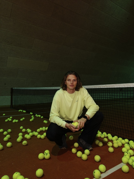 En person sidder foran et net på en indendørs tennisbane, omgivet af mange gule tennisbolde, mens de holder én bold i hænderne.